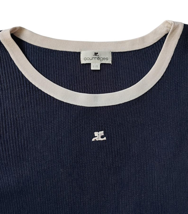 May include: A dark navy blue ribbed knit top featuring a round neckline with a contrasting cream-colored trim. The garment includes a cream-colored embroidered logo on the chest and a white interior label with the text Courr&egrave;ges printed on it.