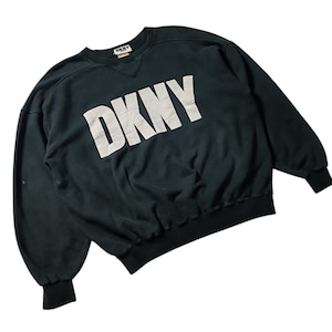 May include: Dark green DKNY sweatshirt with the brand name in large, white, block letters across the front. The sweatshirt has a crew neck, long sleeves, and ribbed cuffs and hem. The DKNY logo is also visible on the neck.