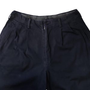 Puede incluir: Primer plano de unos pantalones azul marino. Los pantalones tienen cierre de bot&oacute;n y cremallera, trabillas y pliegues en la parte delantera. La tela parece ser un algod&oacute;n o mezcla resistente. Los pantalones tienen dos bolsillos laterales y un dise&ntilde;o cl&aacute;sico y a medida.