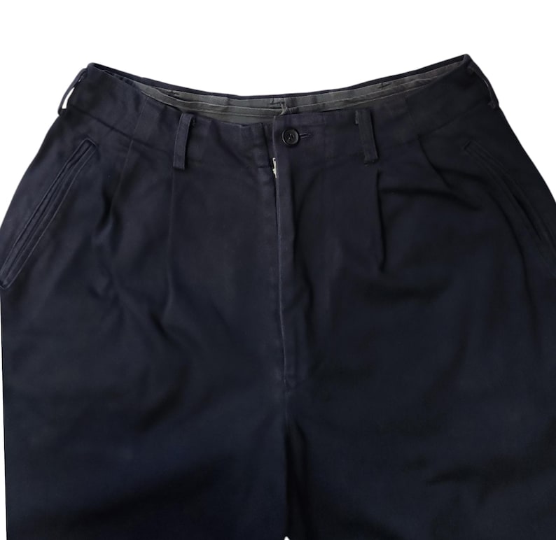 Puede incluir: Primer plano de unos pantalones azul marino. Los pantalones tienen cierre de bot&oacute;n y cremallera, trabillas y pliegues en la parte delantera. La tela parece ser un algod&oacute;n o mezcla resistente. Los pantalones tienen dos bolsillos laterales y un dise&ntilde;o cl&aacute;sico y a medida.