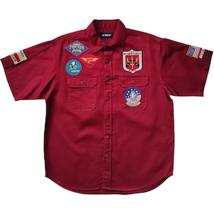 May include: A burgundy short-sleeve shirt with multiple embroidered patches. Patches include text and emblems, such as "TOP TEN", "DALLAS", and a U.S. flag. The shirt has two chest pockets and a button-down front.