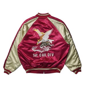 May include: A red satin bomber jacket with a white eagle embroidered on the back. The jacket has a cream colored collar and sleeves. The text "1st CAV. DIV" is embroidered below the eagle.