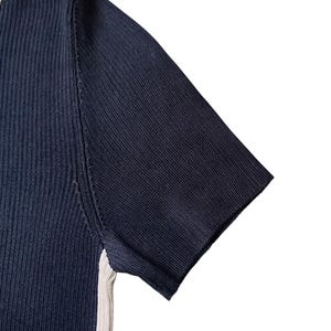 May include: A short-sleeved ribbed knit top in dark navy blue featuring a contrasting cream-colored vertical stripe along the side seam and a matching cream-colored trim along the neckline edge, constructed with a fine vertical texture.