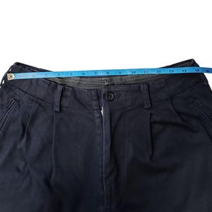 Puede incluir: Primer plano de pantalones azul marino con cierre de bot&oacute;n y pliegues. Una cinta m&eacute;trica azul se extiende por la cintura, indicando una medida de aproximadamente 38 cm.