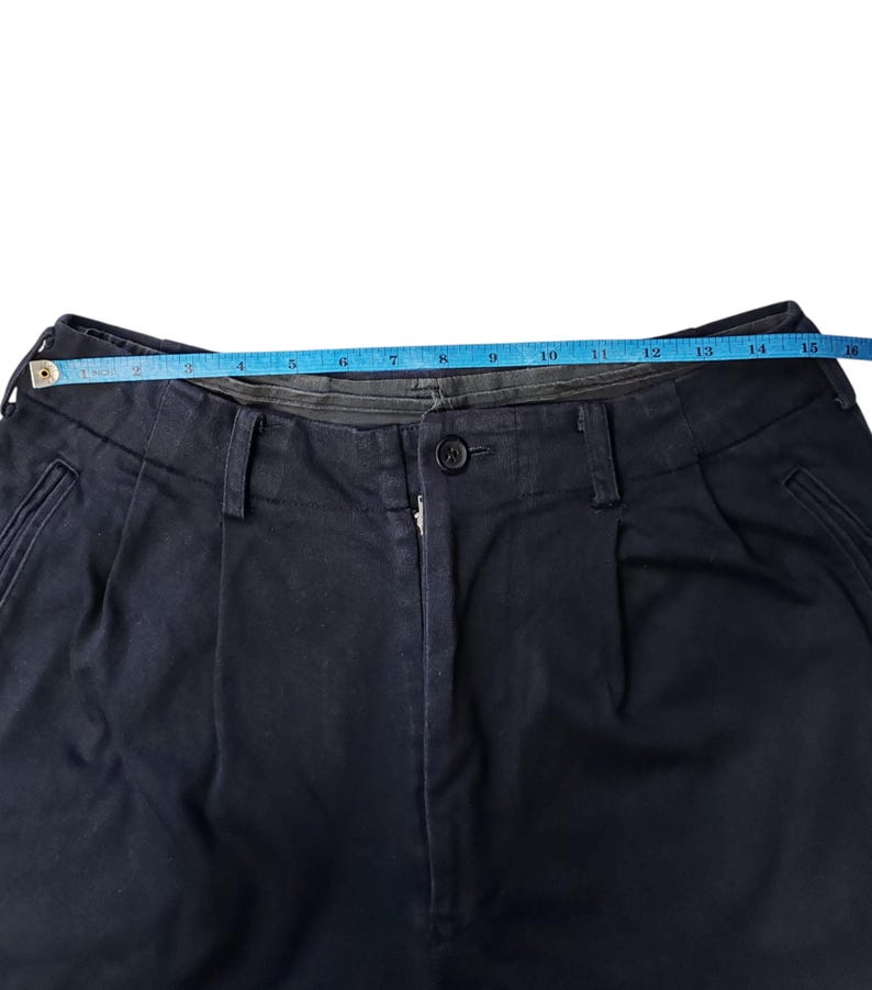 Puede incluir: Primer plano de pantalones azul marino con cierre de bot&oacute;n y pliegues. Una cinta m&eacute;trica azul se extiende por la cintura, indicando una medida de aproximadamente 38 cm.