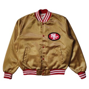 May include: A gold satin bomber jacket with red and white striped trim on the collar, cuffs, and waist. The jacket features a red and black patch with the letters "SF" on the left chest. The jacket has a button-up front.
