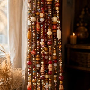 May include: A hanging beaded curtain featuring multiple vertical strands of assorted beads in shades of amber, deep red, brown, and cream. The strands include a mix of faceted glass, smooth wooden, and patterned ceramic beads of various shapes and sizes.