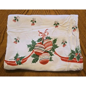 May include: A white fabric tablecloth featuring a repeating pattern of red and green holly sprigs and a central cluster of three red and white striped Christmas ornaments tied with a red ribbon, accented by additional holly and red ribbons.