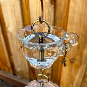 May include: A glass hummingbird feeder with a clear glass cup and a metal key-shaped perch. The feeder is suspended from a metal hook and has a small hummingbird charm hanging from the handle.
