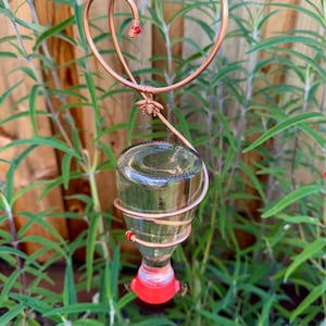 May include: A handcrafted copper hummingbird feeder with a heart-shaped top and a glass bottle. The feeder has a red base and small red beads. A small bee charm is attached to the copper wire. The feeder is hanging among green plants.