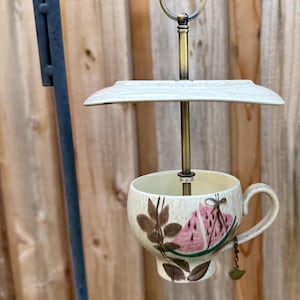 May include: A hanging bird feeder constructed from a cream-colored ceramic teacup and a matching saucer. The cup features a painted design of pink watermelon slices and brown leaves. A brass-toned metal rod connects the pieces with a ring at the top.