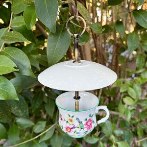 May include: A white porcelain teacup with a floral pattern and a green rim is suspended from a white saucer by a metal rod. The teacup and saucer are attached to a metal hook, which is hanging from a tree branch.