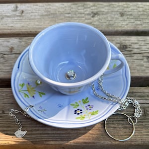 May include: A light blue teacup and saucer set with floral accents. A silver chain with a bird charm and a silver ring are next to the cup. The teacup has a decorative gem in the bottom.