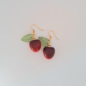 Può includere: Un paio di orecchini pendenti dorati con ciondoli a forma di mela in vetro rosso e accenti in vetro verde a forma di foglia.