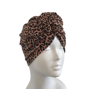 May include: A turban-style head wrap in a leopard print pattern. The fabric features a tan background with brown and black spots. The head covering is knotted at the top and is displayed on a white mannequin head.