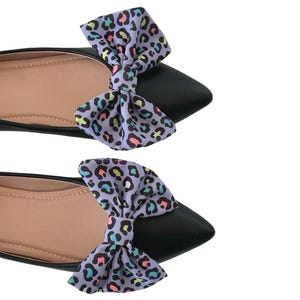 May include: A pair of black pointed-toe flats with a light purple bow featuring a colorful leopard print. The shoes have a neutral-colored insole.