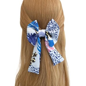 May include: A decorative hair bow featuring a colorful floral and animal print design in shades of blue, white, and pink. The bow is tied and attached to a person's blonde hair, creating a stylish accessory.