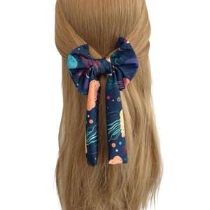 May include: A navy blue hair bow with a celestial pattern, including planets, stars, and colorful nebulas. The bow is tied in the hair, with long, patterned streamers hanging down. The hair is a light brown color.