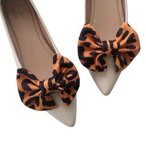 May include: A pair of cream-colored pointed-toe flats with decorative bows. The bows feature a leopard print pattern in shades of orange and black. The shoes are set against a white background.