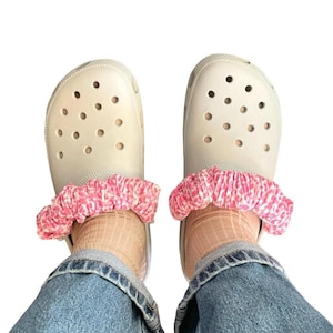 May include: A pair of beige Crocs with pink and white patterned scrunchie-style shoe charms. The shoes are worn with tan socks and blue denim jeans.