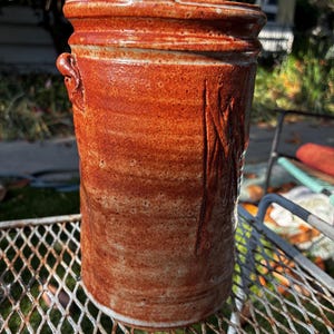 May include: A cylindrical ceramic vessel with a glossy, mottled orange and brown glaze. The exterior features a textured surface with vertical indentations and a small, looped handle near the top rim, showcasing a handcrafted, uneven finish.