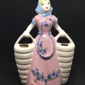 May include: A ceramic figurine of a woman in a pink dress and blue headscarf, holding two white baskets. The dress is decorated with blue and red flowers.