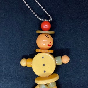 Puede incluir: Un llavero de madera de color amarillo, naranja y verde con forma de persona. El juguete está hecho de cuentas redondas apiladas y tiene una cara pintada con una sonrisa. El llavero está unido a una cadena plateada.