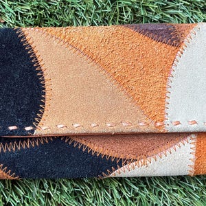 May include: A rectangular wallet featuring a patchwork design of suede leather pieces in black, tan, brown, and cream. The individual sections are joined together with visible, thick orange stitching along the edges of each leather panel.