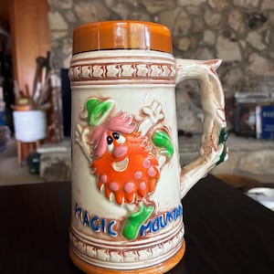 Op de afbeelding: Een keramische bierpul met een reliëf van een kleurrijk stripfiguur met oranje haar en een groene hoed. De tekst "MAGIC MOUNTAIN" staat in blauw aan de basis. De pul heeft een bruin deksel, een getextureerd handvat en decoratieve banden aan de randen.