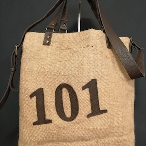 Peut inclure: Un sac cabas en toile de jute naturelle avec des finitions en cuir marron et le numéro "101" imprimé sur le devant.