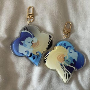 Pode incluir: Dois chaveiros de acrílico transparente com fechos dourados. Cada chaveiro apresenta uma ilustração estilizada de duas figuras em tons de azul, branco e amarelo, sobre um fundo escuro. Os chaveiros têm formato de trevo.