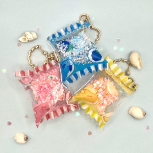 Può includere: Tre portachiavi colorati con glitter all'interno. Ogni portachiavi ha la forma di una piccola borsa con una catena dorata e un ciondolo a forma di conchiglia bianca. Le borse sono trasparenti e mostrano il glitter all'interno. Il glitter è blu, rosa e giallo.