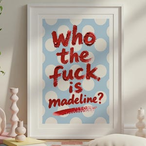 Op de afbeelding: Ingelijste kunstdruk met de tekst "Who the fuck is madeline?" in rode penseelstreken op een lichtblauwe achtergrond met witte stippen. De print zit in een witte lijst en wordt tentoongesteld in een kamer met decoratieve items.