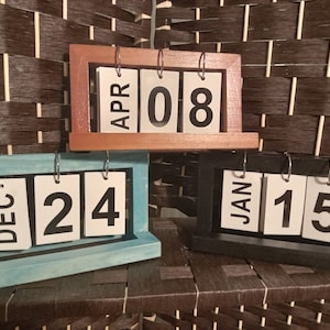 Könnte beinhalten: Drei hölzerne Tischkalender mit umdrehbaren Zahlen und Monaten. Die Kalender sind in verschiedenen Farben: Braun, Blau und Schwarz. Die angezeigten Daten sind APR 08, DEC 24 und JAN 15.