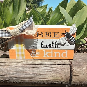 Puede incluir: Un letrero de bloques de madera con las palabras "BEE humble be kind" pintadas en negro y blanco sobre un fondo naranja. Una abeja en blanco y negro está unida a la parte superior del letrero. Una cinta a cuadros en blanco y negro y una cinta de arpillera están atadas alrededor del letrero.