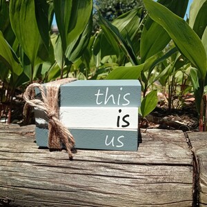 Op de afbeelding: Drie houten blokken gestapeld op elkaar, geschilderd in een lichtgrijze kleur. De blokken hebben de woorden "this", "is" en "us" in witte verf geschreven.