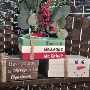 May include: Christmas-themed wooden blocks arranged like books. One stack reads "You're a MEAN ONE Mr. Grinch" in white lettering. Another says "Have yourself a Merry little Christmas". A third stack has a snowman face.