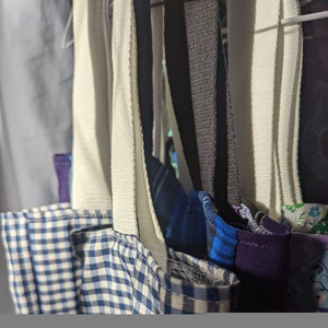 May include: Close-up of several tote bags with different patterned fabric. The bags have white straps and are hanging on a rack.