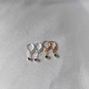 May include: Three pairs of dangle earrings with small gemstone accents. The earrings are silver, gold, and silver with red, green, and green gemstones.
