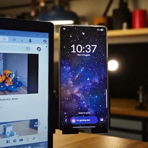 May include: A smartphone displaying a starry sky wallpaper is mounted on a black holder. The screen shows the time 10:37 and the date, Tuesday, August 5. The phone is next to a laptop displaying a webpage.
