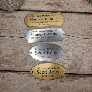 May include: Four gold and silver memorial plaques with engraved text. The top two plaques are rectangular and read "Cherished Memories Of Maureen Battersby ~12th August 1946 - 24th March 2017~ In God's Care." The bottom two plaques are oval and read "In Loving Memory of Scott Roby 1984 - 2022."