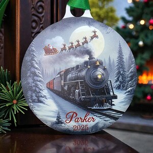 Op de afbeelding: Ronde kerstversiering met een vintage trein op besneeuwde rails, met de slee van de kerstman en rendieren in de lucht. De versiering heeft een groen lint om op te hangen en de naam "Parker" en het jaar "2025" erop gedrukt.