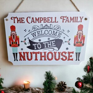 May include: A white wooden sign with the text "THE CAMPBELL FAMILY WELCOME TO THE NUTHOUSE" in red and black. The sign features illustrations of nutcracker soldiers and is suspended by a rope. Christmas decorations are visible around the sign.