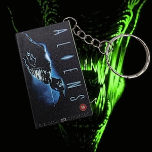 Könnte beinhalten: Ein schwarzer Schlüsselanhänger mit dem Filmtitel "ALIENS" in grüner Schrift. Der Schlüsselanhänger hat ein kleines rechteckiges Design mit einem Filmposterbild und einem silbernen Schlüsselring.