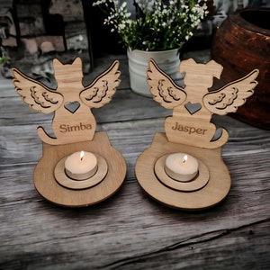 Könnte beinhalten: Zwei Kerzenhalter aus Holz mit Engelsflügeln, einer mit einer Katze und der andere mit einem Hund. Der Katzenhalter ist mit dem Namen "Simba" und der Hundehalter mit dem Namen "Jasper" graviert.