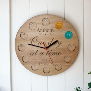 May include: A wooden round clock with a red minute hand and black hour hand. The clock face has a light wood grain and features the text "Anthony One day at a time 12.06.2023". There are 12 small circles around the clock face.