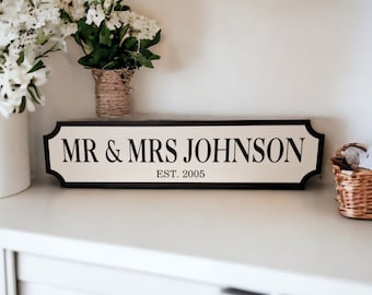 May include: A black and white sign with the text "MR & MRS JOHNSON EST. 2005" on a white background. The sign is on a white shelf with white flowers in a white vase and a wicker basket.