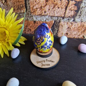 May include: A wooden Easter egg holder with a blue and yellow foil-wrapped chocolate egg on top. The holder has the text "Happy Easter James" engraved on it.