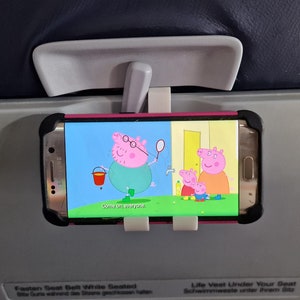 May include: A pink smartphone with a pink case is mounted to the back of an airplane seat. The screen shows a cartoon of a pig with a bucket and a net. The text on the screen reads "Come on everyone".