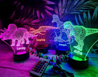 May include: Four 3D printed dinosaur night lights with personalized names: Lacey, Sophie, Joseph, and Alfie. The lights are on a wooden surface and are illuminated in different colors. A remote control with multiple color options is also visible.
